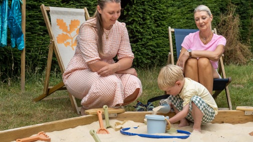 An image of a child playing in a sandpit with two adults sat next to him on deckchairs